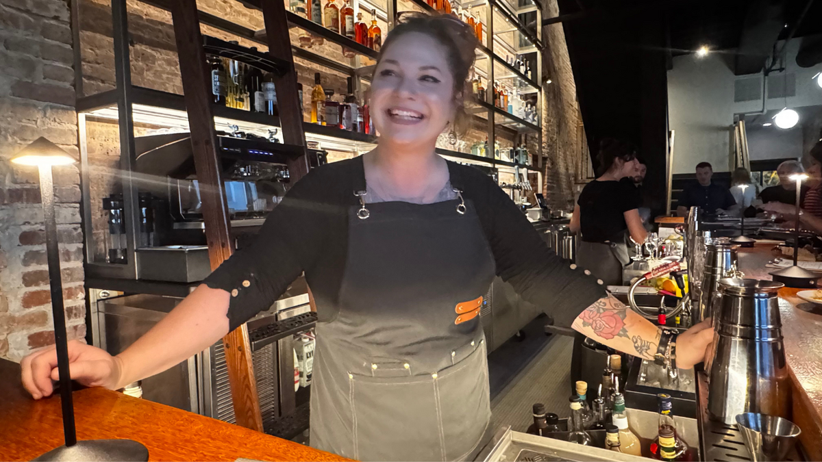 a smiling woman standing behind a cocktail bar